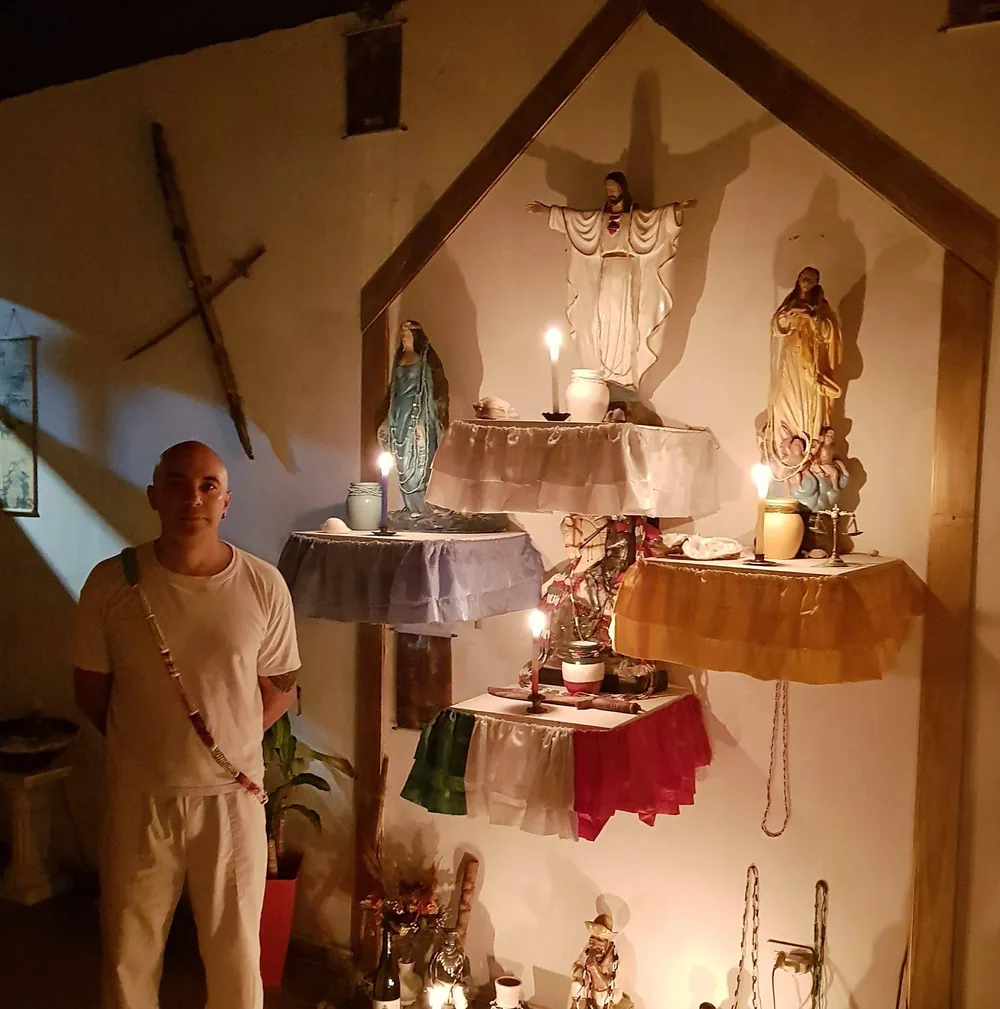 Tayta Miguel de pie junto a su altar ancestral, rodeado de imágenes sagradas y velas encendidas